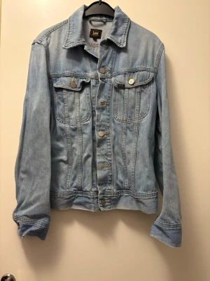 Ljusblå jeansjacka från Lee - Klassisk ljusblå jeansjacka från Lee-slim rider med raka linjer, två bröstfickor med lock och knapp, samt knappar baktill, så den är lite justerbar.