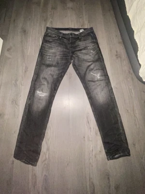 Svarta slitna jeans med raka ben - Säljer ett par svarta jeans med slitningar och distressed detaljer på låren och knäna. Jeansen har raka ben, klassisk femficksmodell och knappgylf. Perfekta för en avslappnad och trendig look.