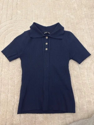Marinblå ribbad polotopp med knappar - Snygg marinblå ribbad polotopp med korta ärmar och fyra knappar framtill. Klassisk krage och stretchigt material som sitter bekvämt. Perfekt till jeans eller kjol för en enkel men stilren look.