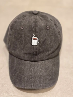 Svart keps med broderad kaffekopp - Snygg svart keps i tvättad bomull med en liten broderad kaffekopp framtill. Justerbar rem med metallspänne baktill för perfekt passform. Perfekt accessoar för dig som älskar kaffe och vill ha en chill stil.