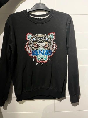 Kenzo sweatshirt - Cool svart sweatshirt från Kenzo med ikoniskt broderat tigerhuvud och färgglada detaljer framtill. Rund halsringning och ribbade muddar vid ärmslut och nederkant. Perfekt statement-plagg för dig som gillar streetstyle.