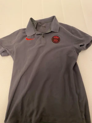 Grå PSG pikétröja från Nike - Snygg grå pikétröja från Nike med Paris Saint-Germain-logga på bröstet och röd Nike swoosh. Tröjan har klassisk krage, korta ärmar och knappar vid halsen. Perfekt för dig som gillar sportig stil och vill representera PSG.