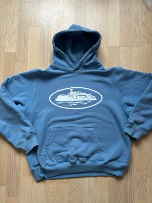 Blå hoodie från Corteiz - Blå hoodie med stor huva och magficka. På bröstet finns ett vitt tryck av en fästning eller byggnad inuti en oval ram. Hoodien har ribbade muddar vid ärmar och nederkant. Perfekt för en avslappnad och cool stil.