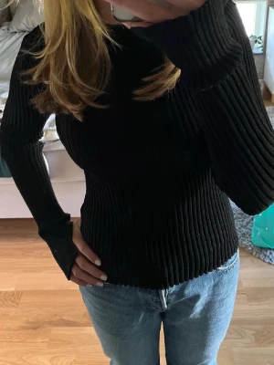 Svart ribbad stickad tröja - Säljer en svart, ribbstickad tröja med lång ärm och hög hals. Tröjan är figurnära och har en stretchig passform som sitter snyggt på kroppen. Perfekt till jeans eller kjol för en clean look. Glitter tröja 