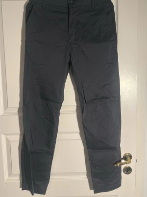 Mörkgrå chinos med rak passform från Nike - Snygga mörkgrå chinos med rak passform och klassiska sidfickor. Byxorna har bälteshällor, knappgylf och två bakfickor med diskreta ficklock. Perfekta för en clean och stilren look. Materialet känns mjukt och lätt, troligen bomull eller bomullsblandning.