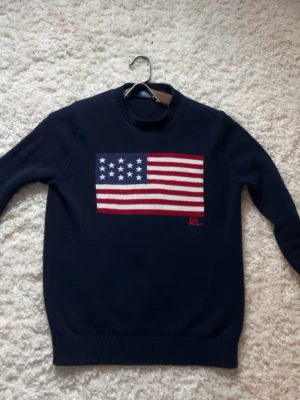 Ralph Lauren tröja - Mörkblå stickad tröja från Ralph Lauren med amerikansk flagga i rött, vitt och blått på bröstet. Rund halsringning och ribbstickade muddar vid ärmslut och nederkant. RL-broderi i rött nedanför flaggan.