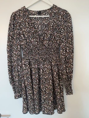 Blommig kortklänning med v-ringning - Söt kortklänning med småblommigt mönster i svart, vit och rosa. Klänningen har v-ringning, långa ärmar och smock i midjan för en snygg passform.