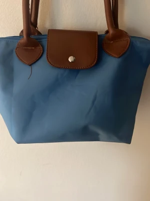 Blå handväska med bruna detaljer - Snygg blå handväska med bruna handtag och lock. Väskan har en klassisk, enkel design med silverfärgad knappstängning och är tillverkad i ett tåligt material som liknar nylon. Perfekt storlek för det viktigaste och lätt att bära över axeln.