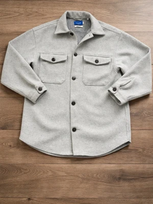 Ljusgrå overshirt från Jack & Jones - Stilren ljusgrå overshirt från Jack & Jones Originals med två bröstfickor med lock och knappar. Klassisk krage, svarta knappar framtill och vid ärmslut. Mjuk och lite tjockare material som passar perfekt som lager på lager-plagg.