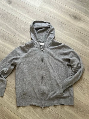 Grå zip från sayless  - En enkel och stilren grå hoodie med dragkedja framtill och två fickor. Tillverkad i mjuk bomullsblandning och har ribbade muddar vid ärmslut och nederkant. Perfekt för en avslappnad look och har en klassisk huva. Tröjan har ett litet hål på ena armbågen, men går lätt att laga.                         Hör av er vid funderingar
