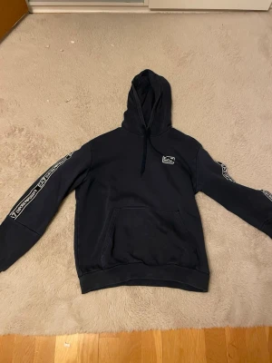 Svart EA7 hoodie med logga - Snygg svart hoodie från EA7 med huva och känguruficka. EA7-logga på bröstet och vita detaljer längs ärmarna med text. Tillverkad i mjukt bomullsmaterial som känns skönt mot huden. Perfekt för en avslappnad och trendig look.