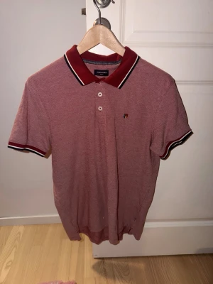 Röd pikétröja från Paul & Shark - Klassisk röd pikétröja från Paul & Shark med vit och svart rand på krage och ärmslut. Tröjan har korta ärmar, knappslå med tre knappar och en liten logga på bröstet. Perfekt till jeans eller shorts för en clean look.