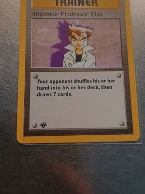 Impostor Professor Oak 1st Edition  - Säljer ett original Pokémonkort från 1999, Impostor Professor Oak. Kortet är i klassisk rektangulär form med gul ram och motiv av Professor Oak. Texten är på engelska och kortet har en distinkt baksida med Pokéball och blå bakgrund.