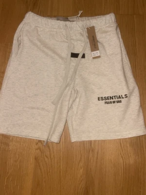Essentials Fear of God shorts ljusgrå - Säljer ett par ljusgrå shorts från Essentials Fear of God med svart logga på vänster ben. Shortsen har snörning i midjan och sidofickor. Perfekta för sommaren och riktigt sköna med sin mjuka bomullskänsla.