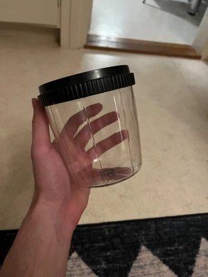 Transparent plastburk med svart lock - Säljer en transparent plastburk med svart skruvlock. Perfekt för förvaring av småsaker, snacks eller andra prylar. Tillverkad i Sverige och har en enkel, stilren design som passar i de flesta hem.