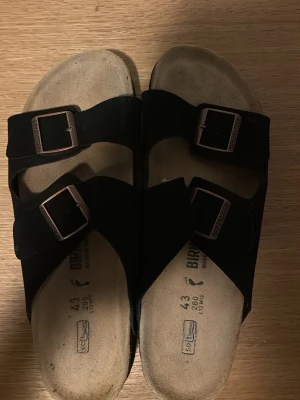 Svarta Birkenstock sandaler - Klassiska svarta Birkenstock sandaler med två breda remmar i mocka och justerbara metallspännen. Fotbädden är ergonomiskt utformad och har en beige färg. Perfekta för en avslappnad och trendig stil.