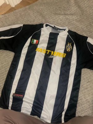 Juventus matchtröja Nedved 11 - Juventus retro matchtröja från Lotto med klassiska svartvita ränder, broderad klubbmärke och italiensk flagga på bröstet. Sponsorlogga FASTWEB i gult framtill och Nedved 11 tryckt i rött på ryggen. Perfekt för fotbollsfans och samlare.