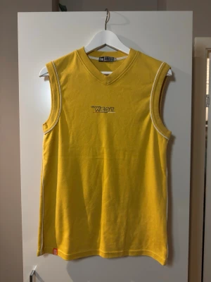 Gul ärmlös linnetopp Go West - Gul ärmlös linnetopp från Go West med broderad logga på bröstet. Toppen har v-ringad hals och är gjord i mjuk bomull. Perfekt för en avslappnad och cool stil.
