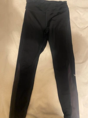 Svarta leggings från Puma med meshdetaljer - Snygga svarta leggings från Puma med diskreta meshpaneler längs benen och en liten logga på vaden. De har hög midja och är tillverkade i ett stretchigt material som sitter skönt och följer kroppen. Perfekta för träning eller chill.