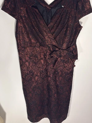 Svart och röd omlottklänning med rosett - Snygg kort omlottklänning i svart och rött med blommigt spetsmönster och dekorativ rosett i midjan. Klänningen har korta ärmar och v-ringning, perfekt för dig som vill sticka ut med en elegant look.