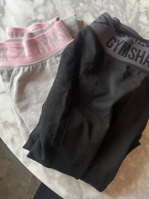 2-pack Gymshark leggings - Två par leggings från Gymshark, ett par i svart med bred midjeresår och logga, och ett par i ljusgrått med rosa midjeresår och logga. .