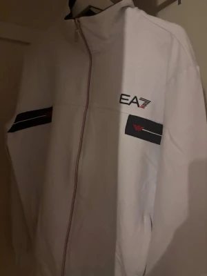 Vit ziptröja med EA7 tryck - Snygg vit ziptröja med EA7 tryck med hög krage och hel dragkedja. Tröjan har svarta detaljer på ärmarna och logga på bröstet samt ärmarna. Perfekt för en sportig och stilren look.
