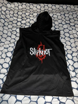 Slipknot ärmlös hoodie svart - Säljer en svart ärmlös hoodie med Slipknot-tryck i vitt och rött framtill. Huvan har dragsko och materialet känns som polyester. Perfekt för dig som vill ha en edgy och unik topp med bandstil.