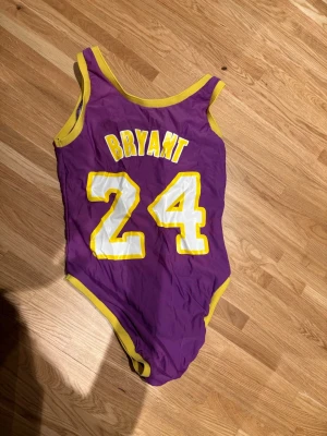 Lila Lakers baddräkt Bryant 24 - Snygg lila baddräkt inspirerad av Lakers med gula och vita detaljer. På framsidan står det 'Lakers des 24' och på baksidan 'Bryant 24'. Klassisk sportig look med breda axelband- perfekt för dig som älskar basket och vill sticka ut på stranden. Finns ingen storlek men skulle säga att den skulle passa s-m