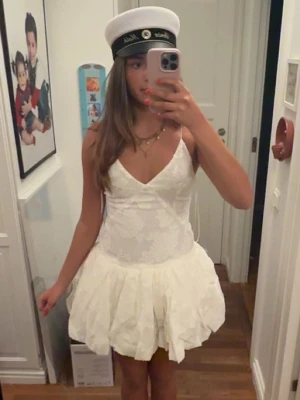 Student klänning  - Säljer en vit kort klänning med spetsdetaljer och v-ringad front. Klänningen är ärmlös med tunna axelband och har en volangig kjol som ger ett lekfullt intryck. Perfekt för student eller sommarens festligheter.