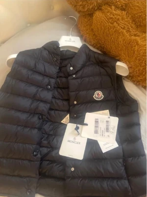 Svart dunväst från Moncler - Snygg svart dunväst från Moncler med klassisk quiltad design och Moncler-logga på bröstet. Västen har tryckknappar framtill och står för både stil och funktion. Perfekt att slänga över en hoodie eller tröja för en trendig look.!!!!Helt ny aldrig använd!!!!!