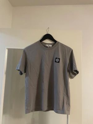 Grå t-shirt från Stone Island - Snygg grå t-shirt från Stone Island med klassisk rund hals och korta ärmar. På bröstet sitter en svart patch med Stone Island-loggan. Tillverkad i mjuk bomull som känns skön mot huden. Perfekt för dig som gillar stilrena och trendiga plagg. Den är väldigt ny har haft den på mig ett par få gånger säljs eftersom den är liten