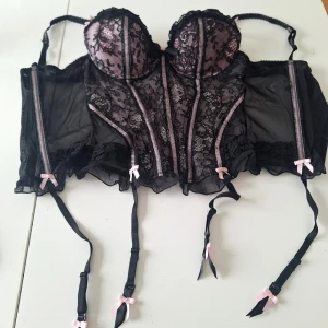 Vintage korsett svart/rosa - Snygg korsett i svart och rosa med vadderade kupor och dekorativ spetsdetalj framtill. Axelbanden är justerbara och sidorna är i genomskinligt meshmaterial. Kuporna har ett diskret mönster och korsetten ger en feminin siluett. Många hyskor bak, därför passar den många. Använd 1 g. Köpt för 25 år sedan ca. 