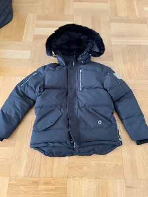 Svart pufferjacka från Moose Knuckles - Säljer en svart pufferjacka från Moose Knuckles med avtagbar huva och fuskpäls. Jackan har flera fickor med dragkedja och tryckknappar samt den ikoniska loggan på ärmen. Perfekt för kalla vinterdagar och riktigt snygg streetstil.