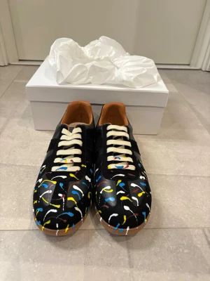 Svarta sneakers med färgstänk - Unika svarta sneakers med färgstänk i blått, gult, vitt och rött. Skorna har vita snören, bruna detaljer på insidan och klassisk gummisula.Materialet är slätt läder och designen är lågskuren.