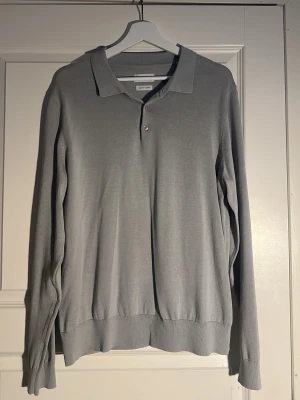 Ljusgrå pikétröja från Urban Quest - Snygg ljusgrå långärmad pikétröja från These glory days. Tröjan har klassisk krage med två knappar framtill och är tillverkad i ett tunt, mjukt material som ger en clean och stilren look. Perfekt för dig som gillar minimalistisk stil.