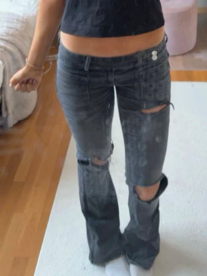 Gina jeans tall - Säljer ett par grå bootcut jeans med slitningar och hål på båda knäna. Jeansen har låg midja, dubbla knappar i linningen och justerbar midja med snöre på insidan.  Strl s tall från Gina 14+ de har en liten fläck där bak men man ser knappt de❤️