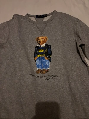 Grå sweatshirt Polo Bear Ralph Lauren - Grå sweatshirt från Ralph Lauren med ikoniska Polo Bear-trycket framtill. Tröjan har rund halsringning och långa ärmar. Perfekt för en avslappnad och trendig stil.