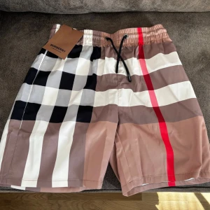 Burberry rutiga shorts med snörning - Snygga shorts från Burberry med klassiskt rutigt mönster i beige, svart, vitt och rött. Shortsen har elastisk midja med svart snörning och är perfekta för sommaren. Ikonisk design som sticker ut och passar till många olika stilar.