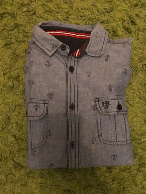 Blå kortärmad skjorta med ankare - Snygg blå skjorta från HR med korta ärmar och små ankare som mönster. Skjortan har två bröstfickor med knappar och klassisk krage. Perfekt för en avslappnad stil och tillverkad i mjuk bomull.