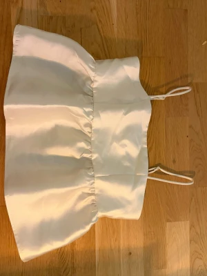 Vit satinlinne med tunna axelband - Snyggt vitt satinlinne med tunna axelband och lätt puffig form. Toppen har smock i ryggen för skön passform och en glansig finish som ger en lyxig känsla. Perfekt att styla med jeans eller kjol för en trendig look.