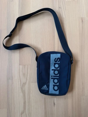 Mörkblå axelväska från Adidas - Snygg mörkblå axelväska från Adidas med justerbar rem och stort logotryck i svart på framsidan. Väskan har en kompakt, rektangulär form och är tillverkad i slitstarkt tygmaterial. Perfekt för att bära det viktigaste och ger en sportig vibe.