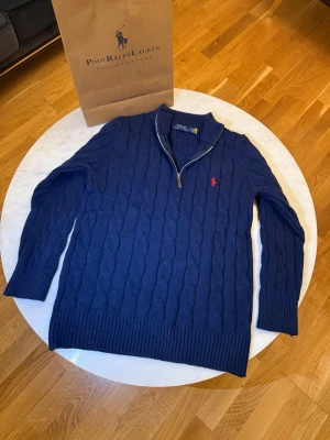 Blå kabelstickad tröja från Polo Ralph Lauren - Snygg blå kabelstickad tröja från Polo Ralph Lauren med halv dragkedja och ribbade muddar. Tröjan har ett klassiskt rött broderat logotyp på bröstet och hög krage. Perfekt för dig som vill ha en stilren och tidlös look.