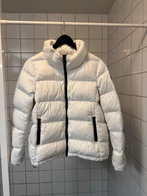 Vit pufferjacka från Colmar - Snygg vit pufferjacka från Colmar med hög krage och svarta dragkedjor framtill och vid fickorna. Jackan har ett stilrent, quiltat mönster och Colmar-logga på ärmen. Perfekt för kalla vinterdagar och ger en clean look.