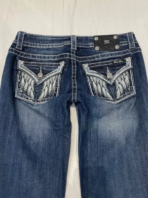 Blå Miss Me jeans med broderade fickor - Snygga blå jeans från Miss Me med raka ben och coola broderade bakfickor i silver och vitt. Jeansen har dekorativa knappar och detaljer på fickorna samt klassisk denimtvätt. Perfekta för dig som vill sticka ut med din stil.