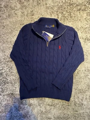 Marinblå Ralph Lauren quarter-zip - En elegant kabelstickad quarter-zip i blått med röd logga. Levererar behaglig upplevelse på, och en stilren look samtidigt. Storleken är M fast passar som en mindre S enligt mig, skicket är mycket bra eftersom den är näst intill oanvänd. Hör gärna av dig vid funderingar så löser vi det! 😁