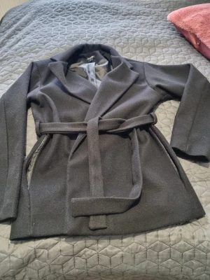 Svart kappa med knytskärp från boohoo  - Stilren svart kappa från boohoo med klassisk krage och knytskärp i midjan. Kappan har fickor på sidorna  Perfekt för dig som vill ha en enkel men snygg look till hösten eller vintern. Storlek 38 men passar Storlek 40 också den är stor i storleken. På bilden ser ljusare ut på grund av kameran. Använd fåtal gånger. 