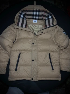 Beige dunväst med huva från Burberry - Stilren beige dunväst från Burberry med avtagbara ärmar och klassiskt rutigt foder i huvan. Västen har två svarta dragkedjefickor framtill och stängs med både dragkedja och knappar. Perfekt för lager-på-lager och riktigt snygg till höst och vinter.