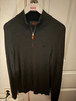 Mörkgrå halvzip-tröja Morris - Snygg mörkgrå stickad tröja från Morris med halv dragkedja och liten logga på bröstet. Tröjan har hög krage och ribbade muddar vid ärmslut och nederkant. Perfekt för lager på lager och stilrena outfits.
