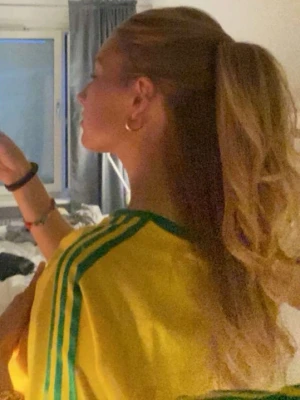 Gul Adidas t-shirt med gröna detaljer - Gul t-shirt från Adidas med gröna ränder på axlarna och gröna muddar vid ärmar och hals. Klassisk Adidas-logga i blått på bröstet. Den är inte lika ”skrikgul” som på bilderna från nätet. Mina egna bilder är hur den är i verkligheten. 