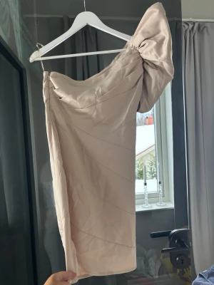 Champagnefärgad cockltailklännkng  - Snygg kort klänning i champagnefärg med en stor puffärm på ena axeln. Klänningen har en asymmetrisk one shoulder-design och är tillverkad i ett glansigt material som ger en lyxig känsla. 